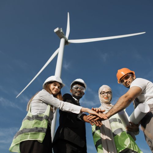 Group of four multicultural partners stacking hands together and smiling on camera while standing among wind turbines. Men and women in safety helmets having successful meeting outdoors.